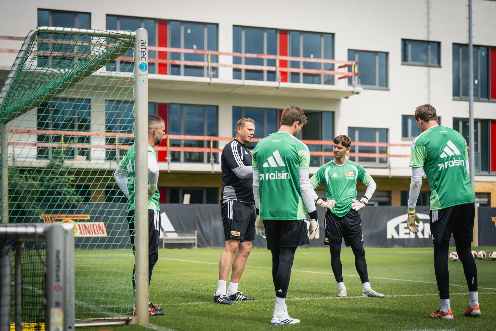 Fußballtorwarttraining mit mehreren Spielern in grünen Trikots auf einem Rasenplatz vor modernen Gebäuden.