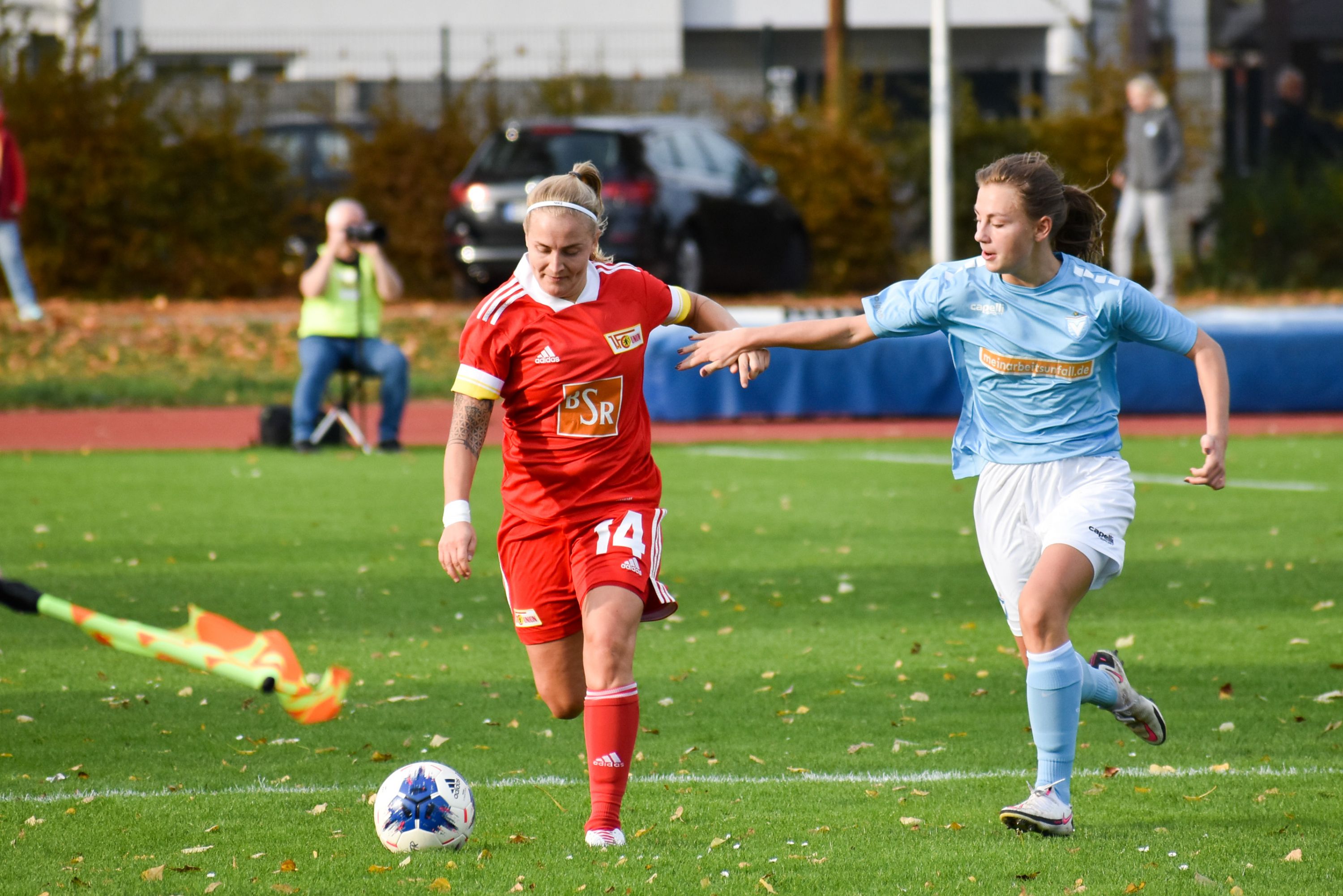 Spielszene eines Fußballspiels, zwei Spielerinnen in roten und blauen Trikots jagen den Ball auf einem grünen Rasenfeld.