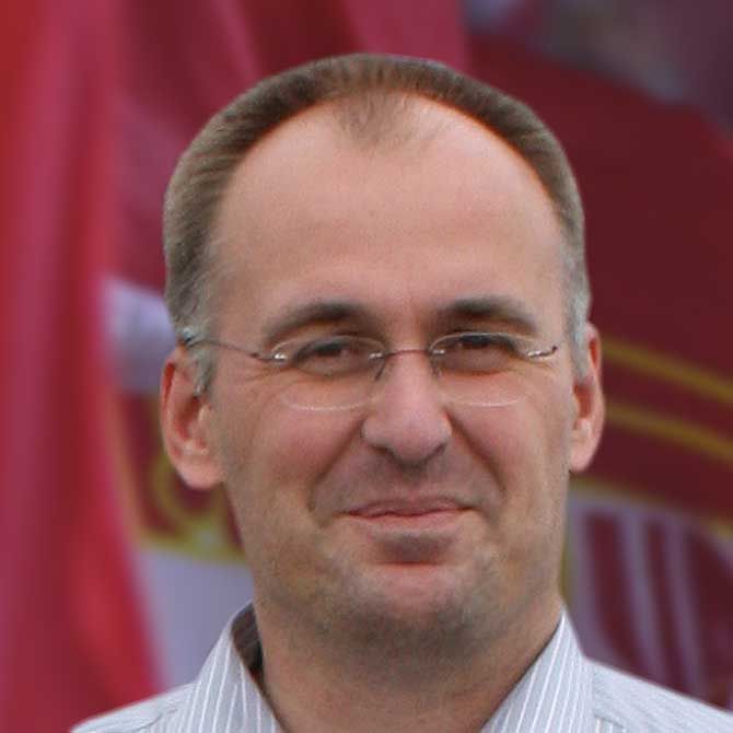 Ein Mann mit Brille und kurzen Haaren lächelt vor einem verschwommenen, rot-weißen Hintergrund.