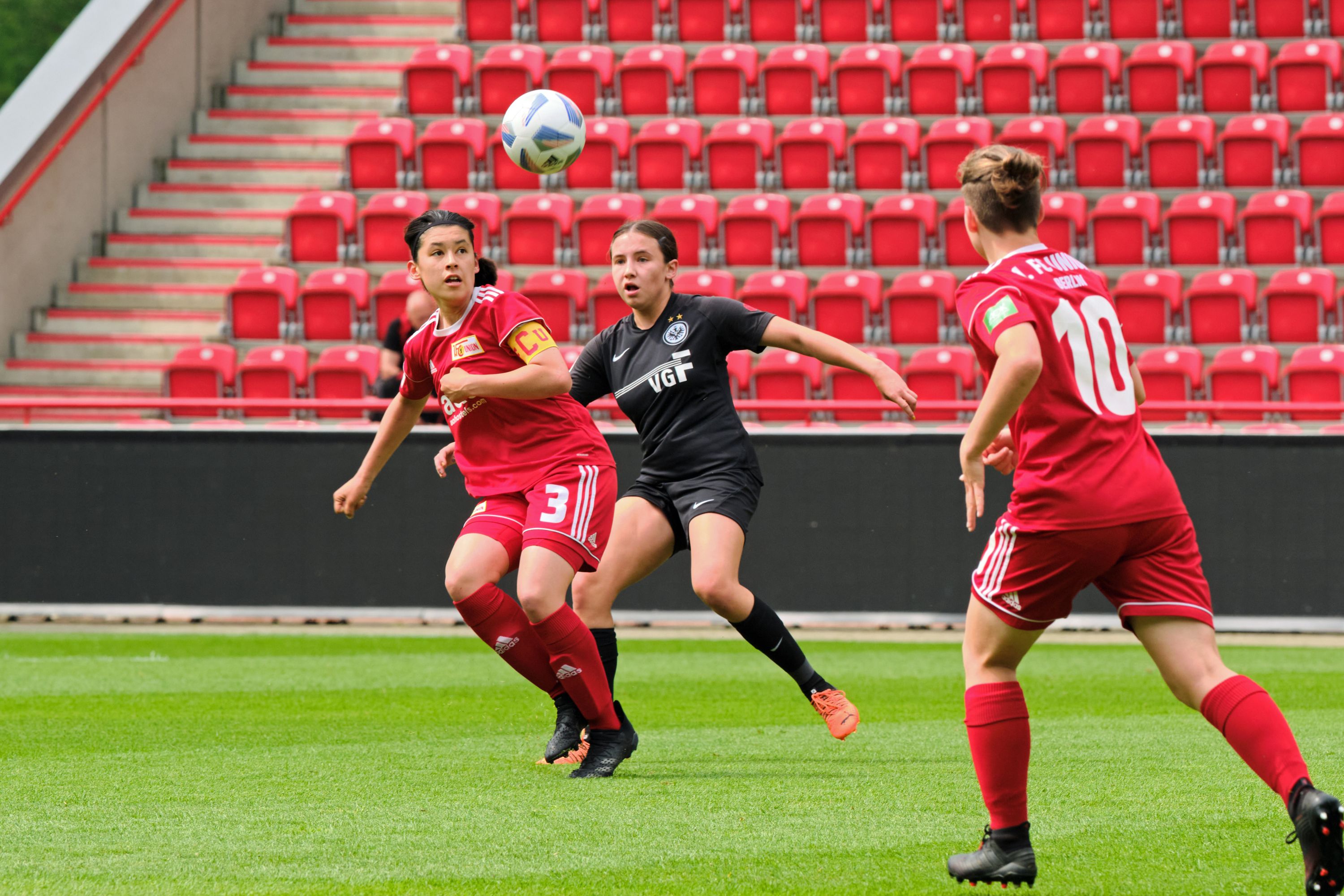 Spielszene im Fußball mit zwei Spielerinnen in roten Trikots und einer Torhüterin in Schwarz, die einen Ball abfängt.
