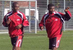 Zwei Fußballspieler in roten Trainingsanzügen laufen auf einem Sportplatz.