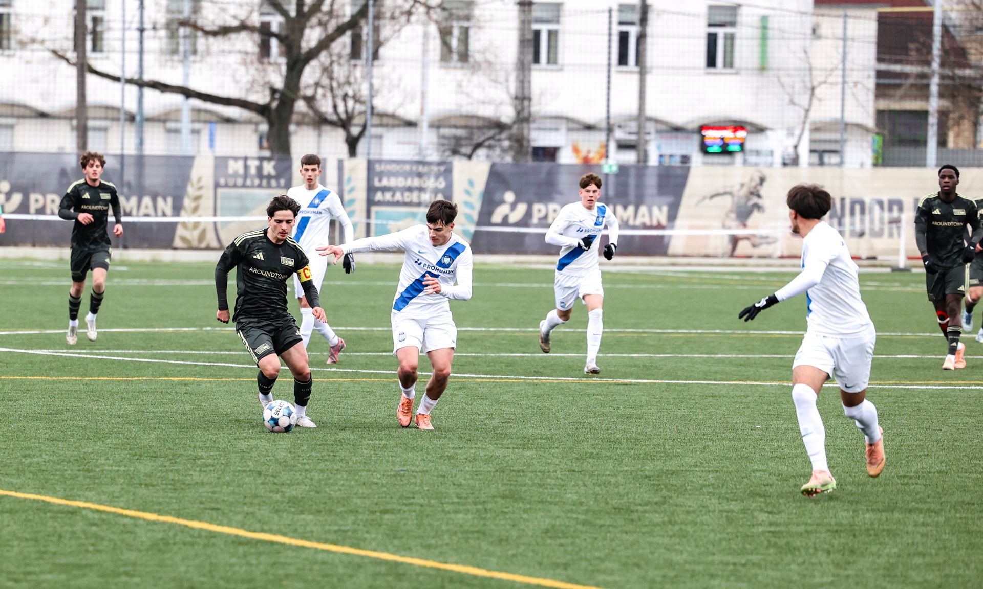 Ein Fußballspiel auf einem Kunstrasenplatz mit mehreren Spielern, die um den Ball kämpfen. Einige Spieler tragen weiße und andere schwarze Trikots.
