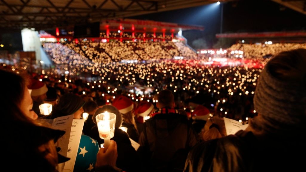Crowd in an arena at night holding candles and singing, surrounded by festive lighting.