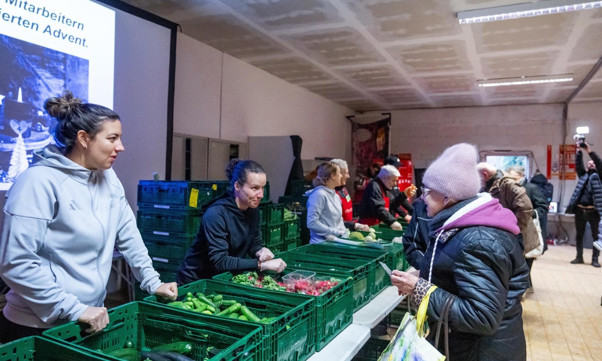 Freiwillige packen frisches Gemüse in Kisten für eine Veranstaltung, während eine Frau mit einer Tasche beobachtet.