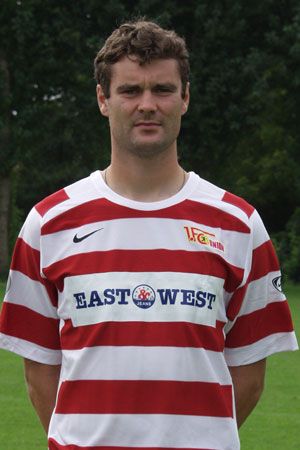 Ein Mann steht in einem gestreiften Sporttrikot mit dem Aufdruck "EAST WEST" und einem Vereinslogo, vor einem grünen Hintergrund.