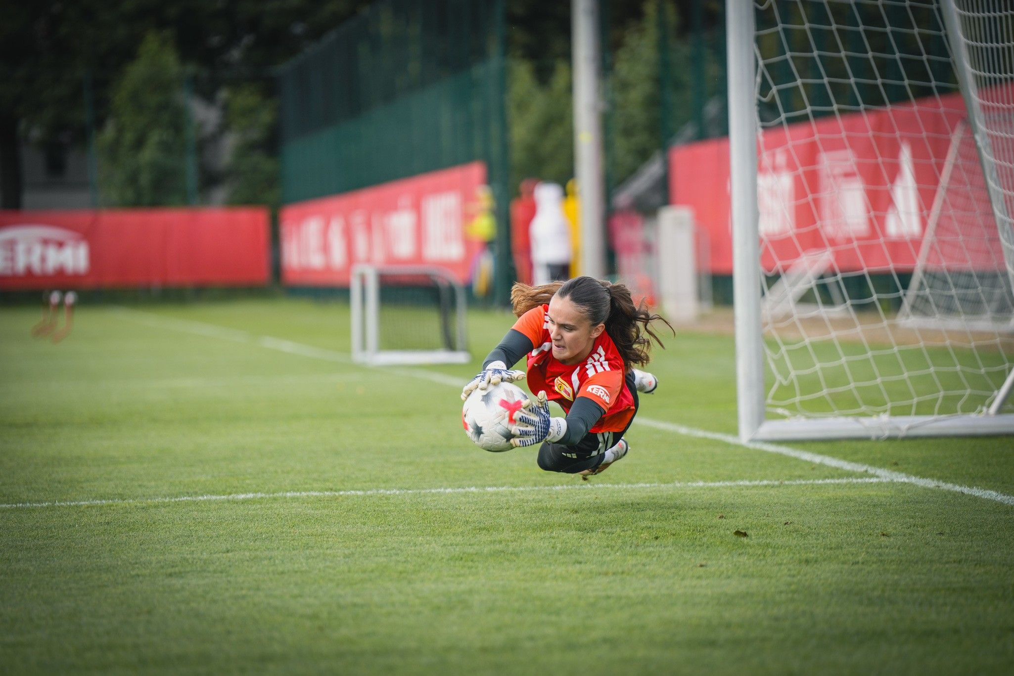 Torhüterin springt im Gras, um einen Fußball während des Trainings zu fangen, mit einem Tor im Hintergrund.