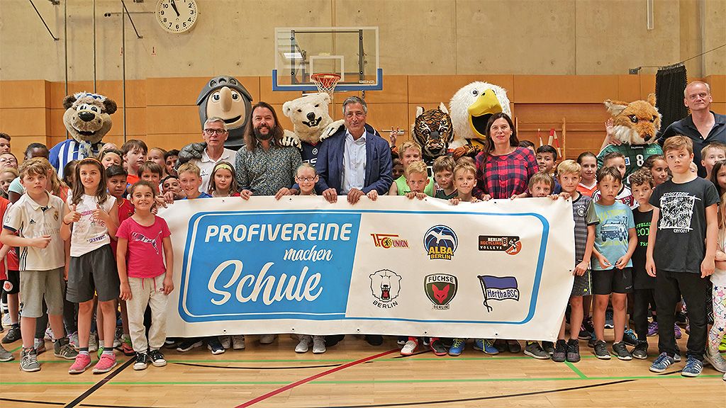 Kinder und Erwachsene posieren in einer Sporthalle vor einem Banner mit der Aufschrift "Profivereine machen Schule", umgeben von Maskottchen.