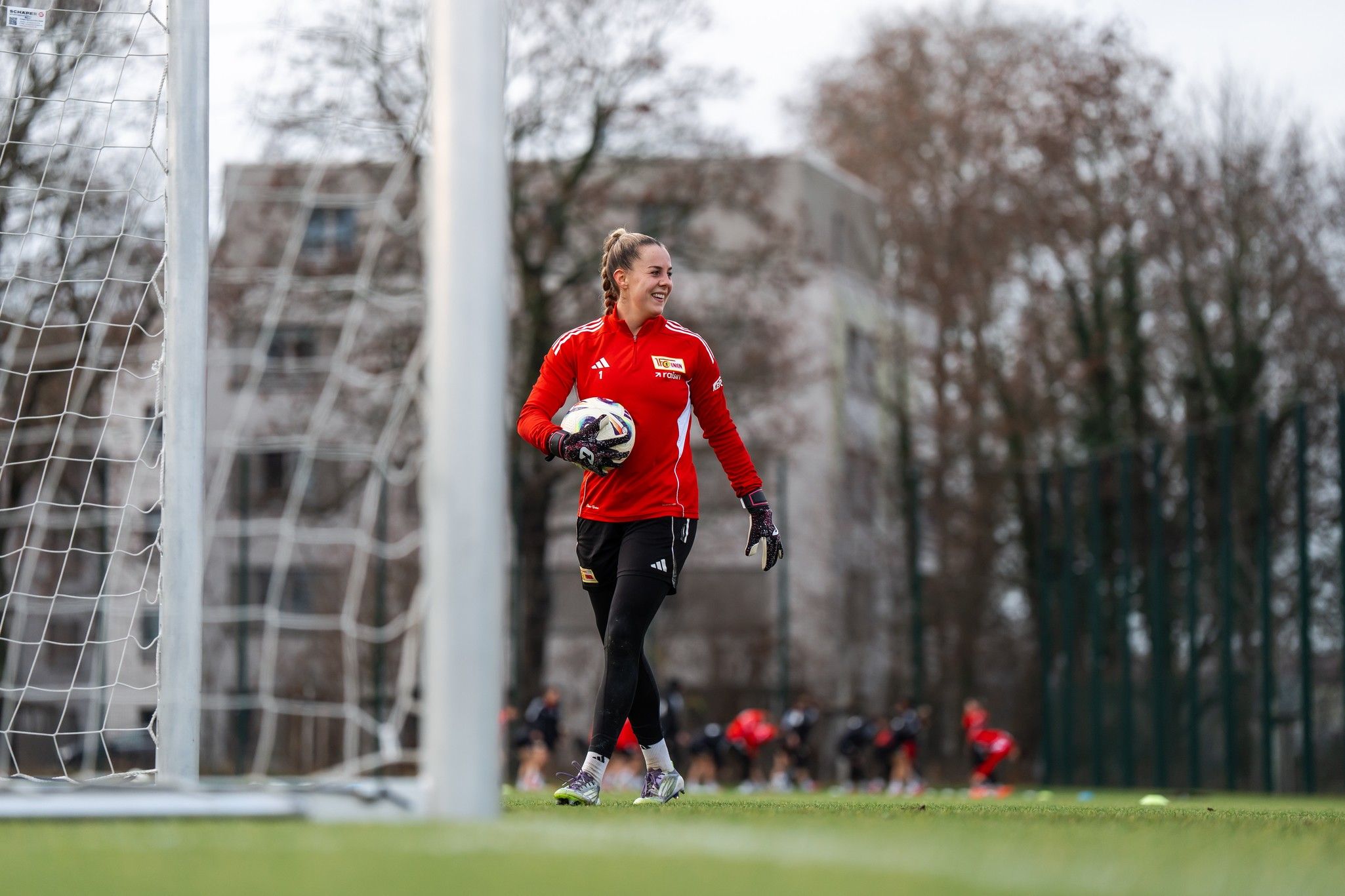 Eine Torhüterin in rotem Trikot hält den Ball und lächelt auf einem Fußballfeld. Im Hintergrund sind Spielerinnen zu sehen.