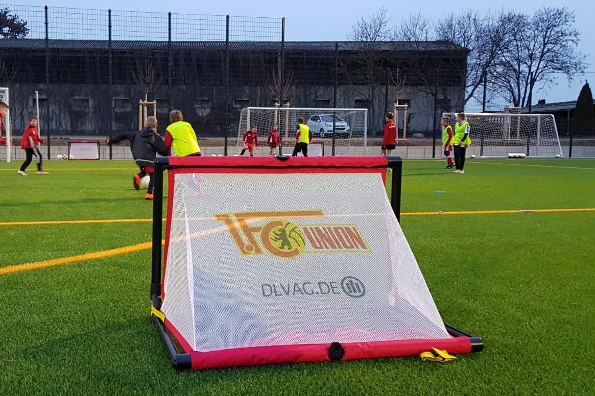 Tor auf einem Fußballplatz mit Spielern im Hintergrund, die trainieren. Ein gelbes Vereinslogo ist auf dem Tor zu sehen.