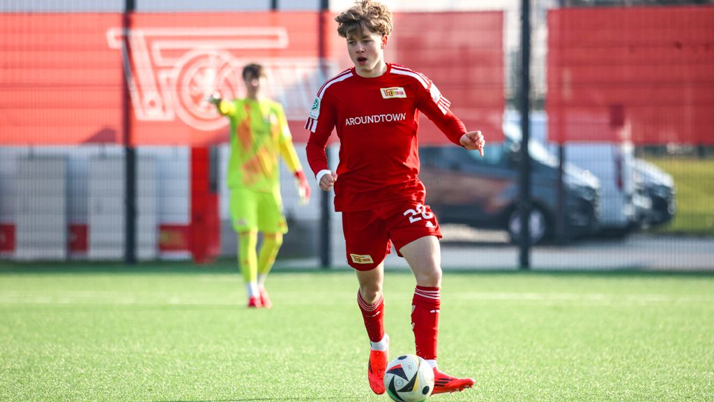 Ein junger Fußballspieler in rotem Trikot dribbelt über ein grünes Spielfeld. Im Hintergrund steht ein weiterer Spieler in gelb.