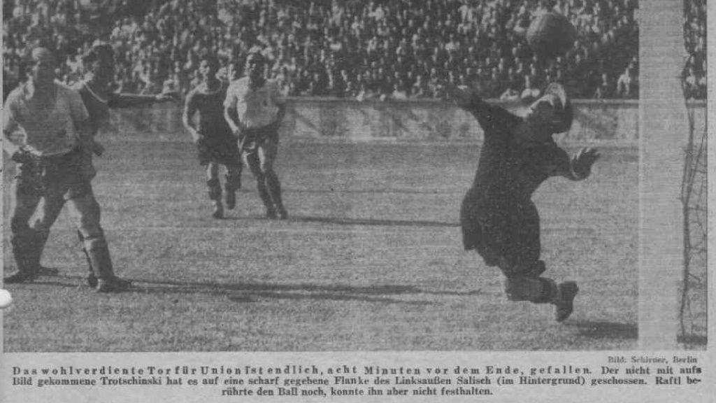 Ein Torwart wehrt einen Ball ab, während Spieler im Hintergrund auf das Tor zielen. Historisches Fußballspiel in Berlin.