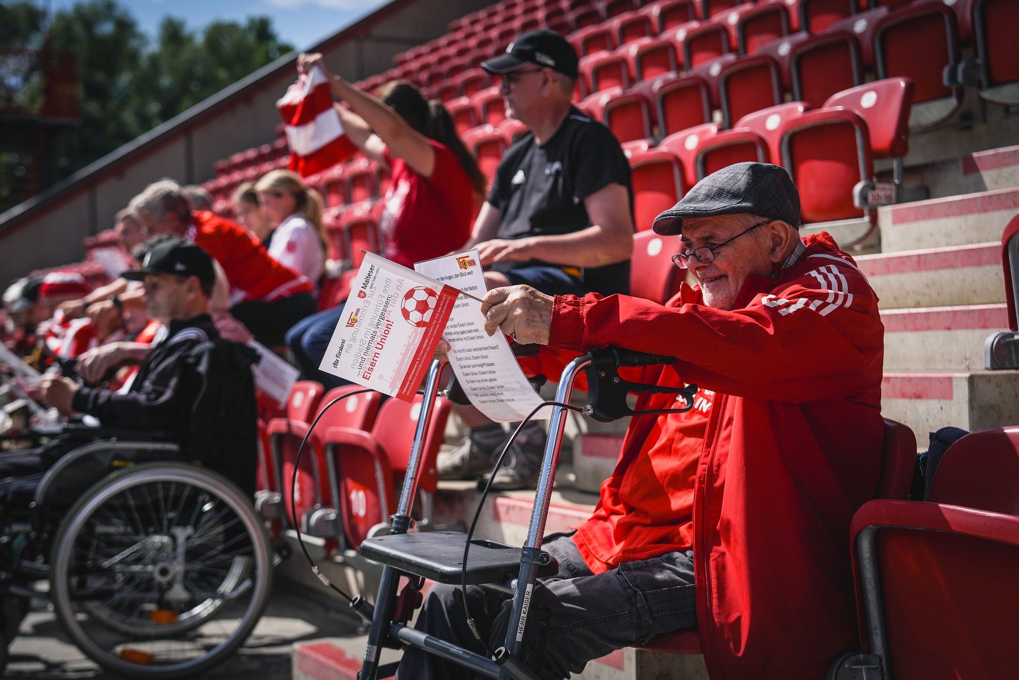 Eine Gruppe von Fans sitzt auf den Rängen eines Stadions, einige halten Fahnen und Plakate, während ein älterer Mann in Rot auf einem Rollator sitzt.