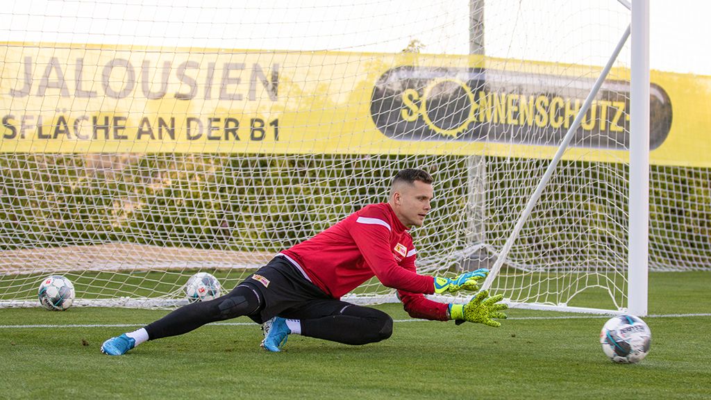 Torwart in roter Trainingsbekleidung dibt sich, um einen Ball im Tor zu fangen, mit einem Netz und weiteren Bällen im Hintergrund.