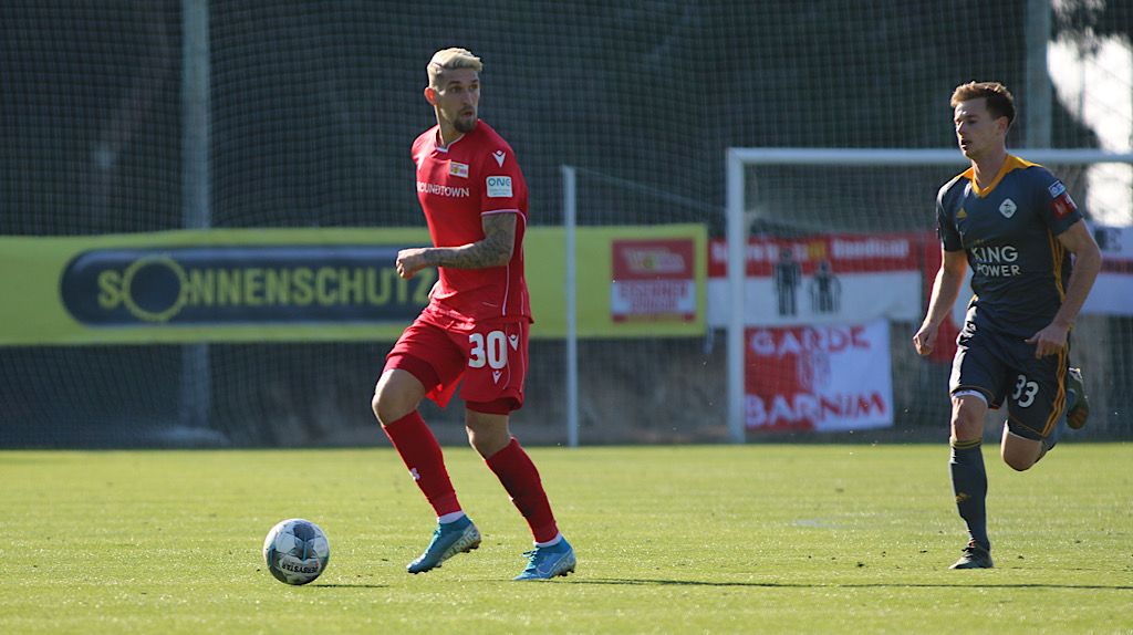 Spielszene mit zwei Fußballspielern auf dem Feld, einer in rotem Trikot mit der Nummer 30 und der andere in einem grauen Trikot mit der Nummer 33.