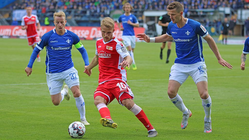 Zwei Spieler in blauen Trikots laufen. Sie folgen einem Spieler in rotem Trikot. Der rote Spieler spielt mit dem Ball einen Pass. Im Hintergrund ist ein Stadion.