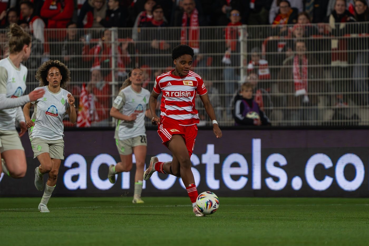 Eine Fußballspielerin in rotem Trikot dribbelt den Ball, während zwei Gegnerinnen in grauen Trikots versuchen, ihn zu verteidigen.