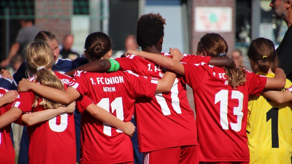 Mädchenmannschaft in roten Trikots umarmt sich vor einem Spiel, Rückenansicht, Nummern sichtbar.