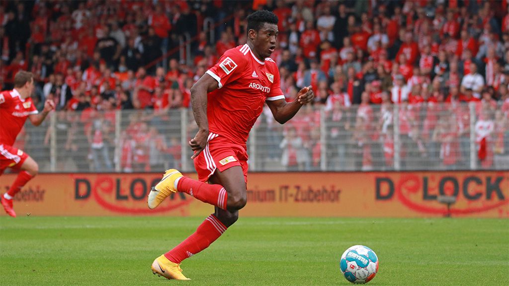 Ein Fußballspieler in rotem Trikot läuft mit dem Ball auf dem Spielfeld, im Hintergrund jubelnde Fans.