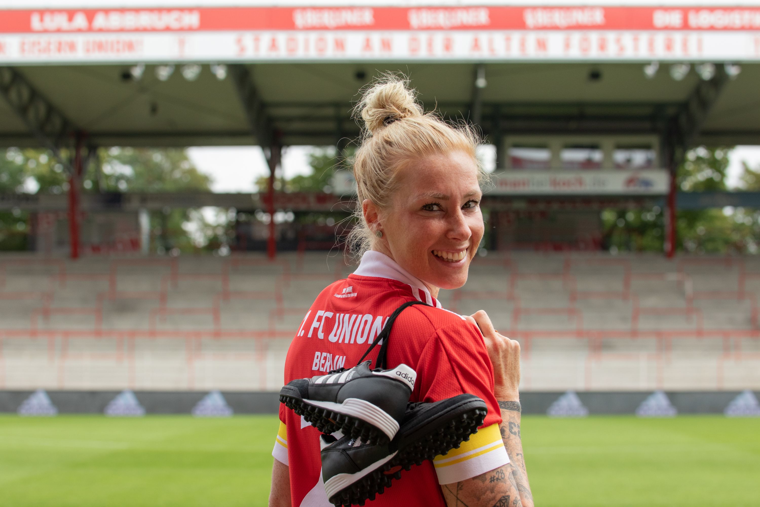 Eine Frau in einem roten Sporttrikot mit der Aufschrift "FC Union Berlin" hält Fußballschuhe über der Schulter auf einem Stadionfeld.