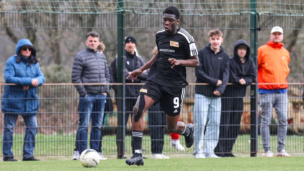 Ein junger Fußballspieler in schwarzer Sportkleidung dribbelt den Ball, während Zuschauer am Spielfeldrand zuschauen.