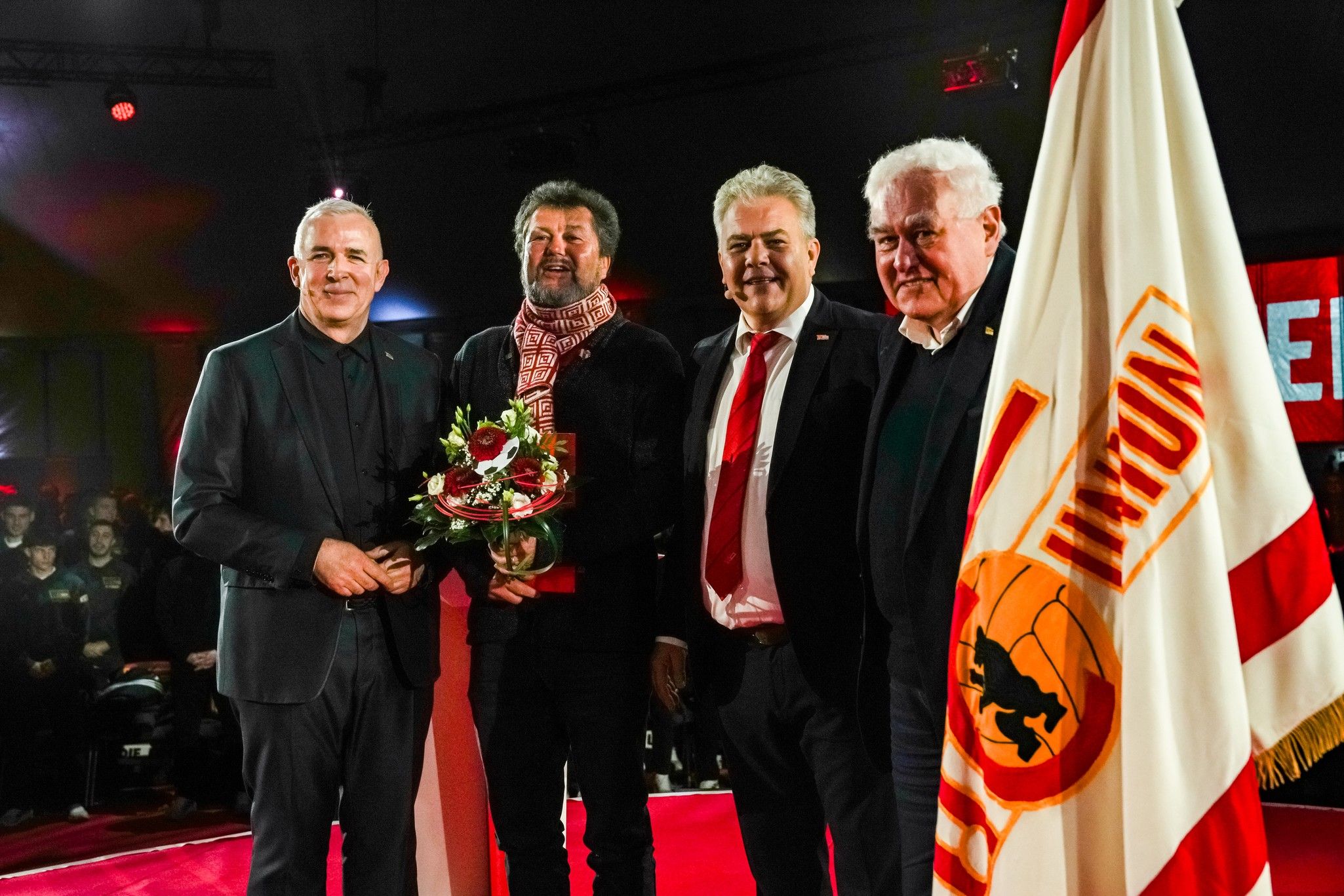 Vier Männer stehen auf einer Bühne, einer hält einen Blumenstrauß. Im Hintergrund sind rote Lichter und eine Fahne zu sehen.