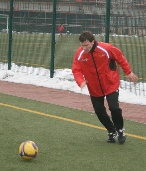 Ein Trainer in roter Jacke spielt außerhalb mit einem Fußball auf einem Trainingsplatz mit grünem Rasen.