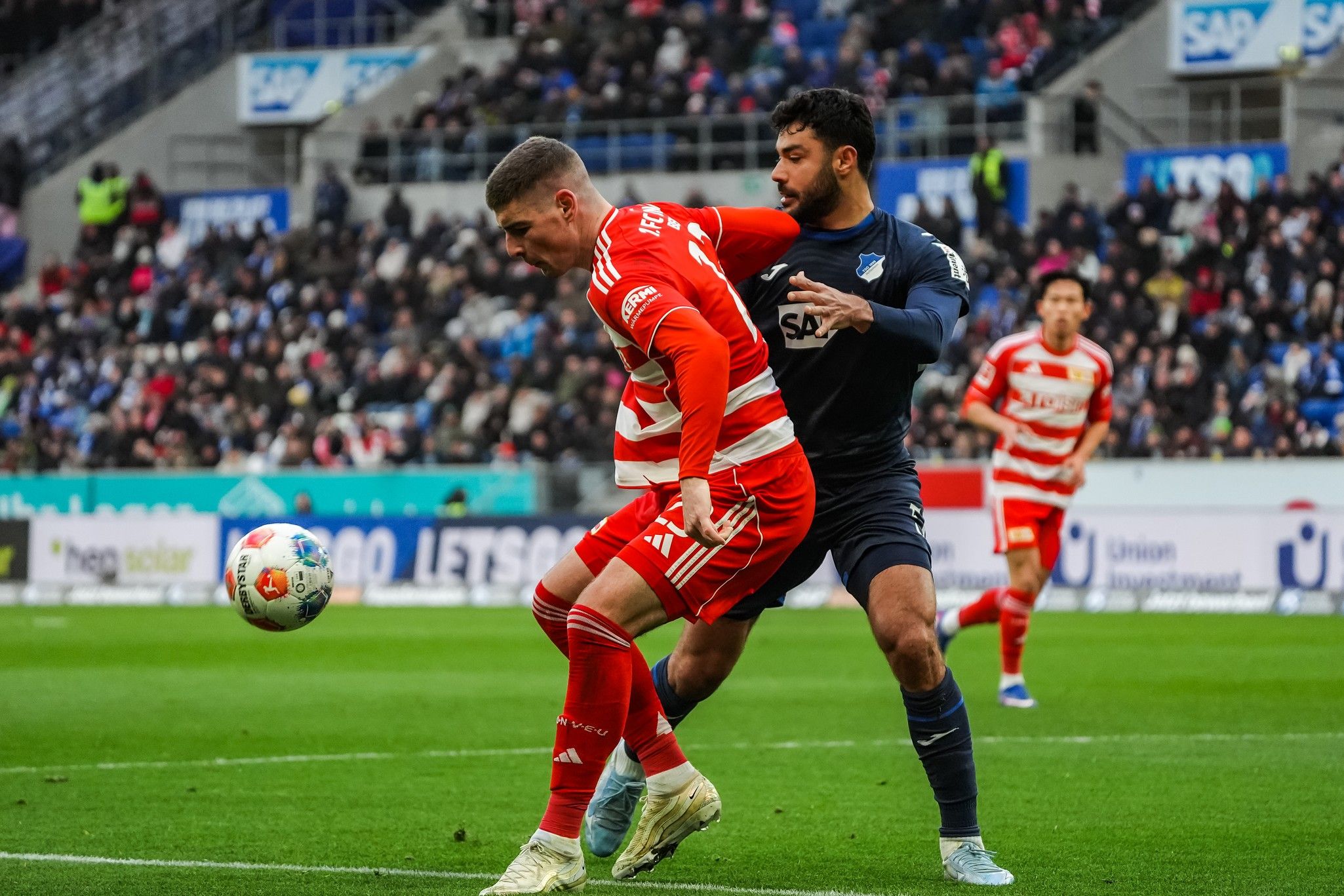Ein Fußballspieler in roter Uniform wird von einem Spieler in blauer Uniform im Wettkampf um den Ball bedrängt.