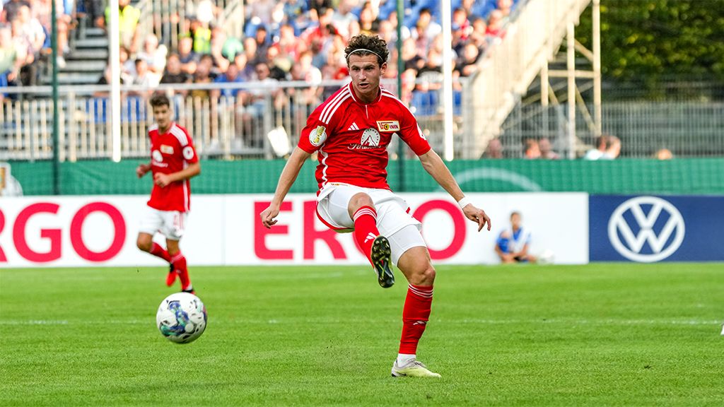 Ein Fußballspieler in rotem Trikot tritt den Ball auf einem grünen Spielfeld während eines Spiels. Zuschauer im Hintergrund.