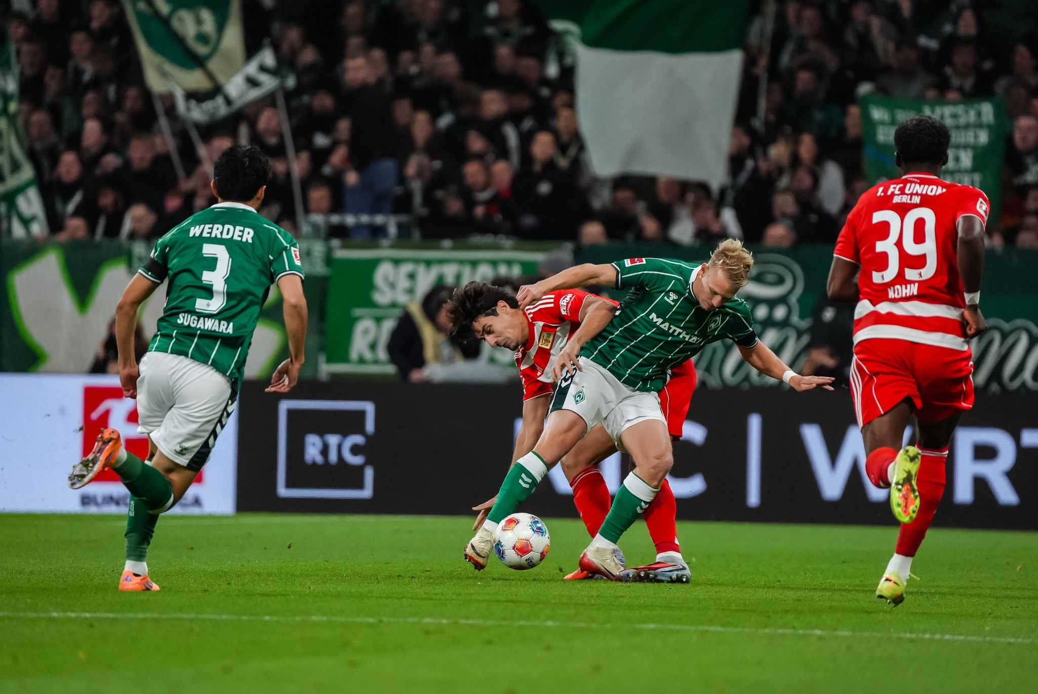 Zwei Fußballspieler kämpfen um den Ball in einem Stadion, während Zuschauer im Hintergrund jubeln.