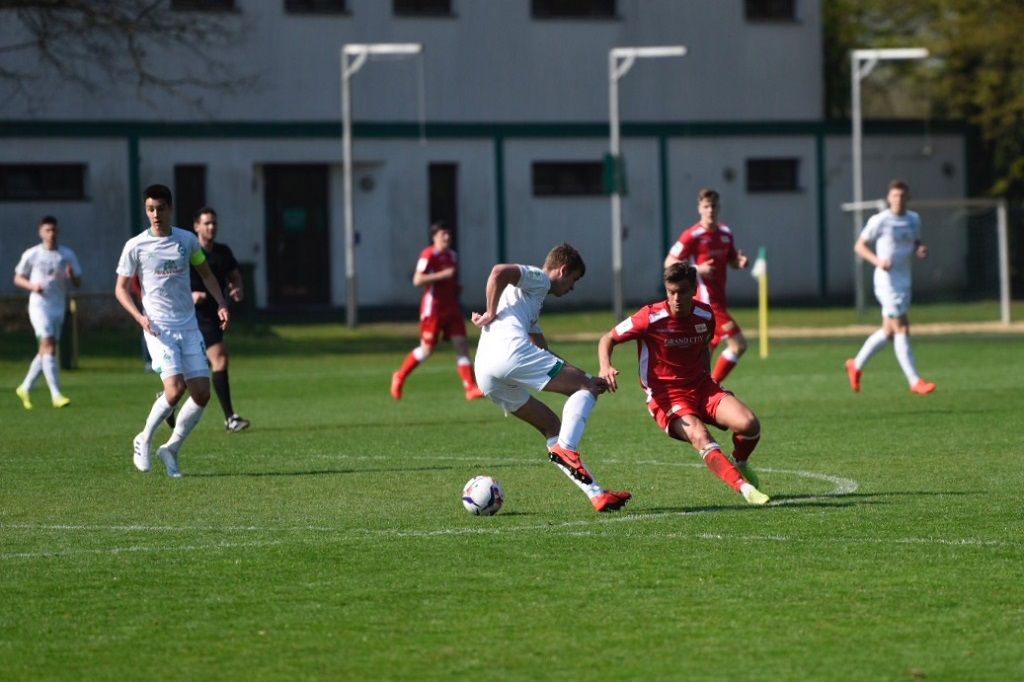 Zwei Fußballspieler kämpfen um den Ball auf einem grünen Spielfeld, während weitere Spieler im Hintergrund stehen.