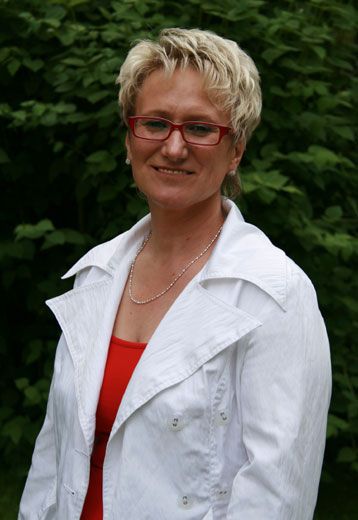 Frau mit blonden, kurzen Haaren und roten Brille, trägt ein weißes Oberteil und steht vor grünem Hintergrund.