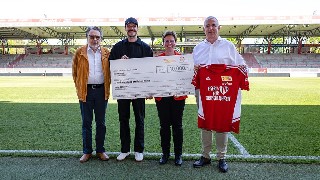 Four people are standing on a soccer field holding a large check and a red jersey.