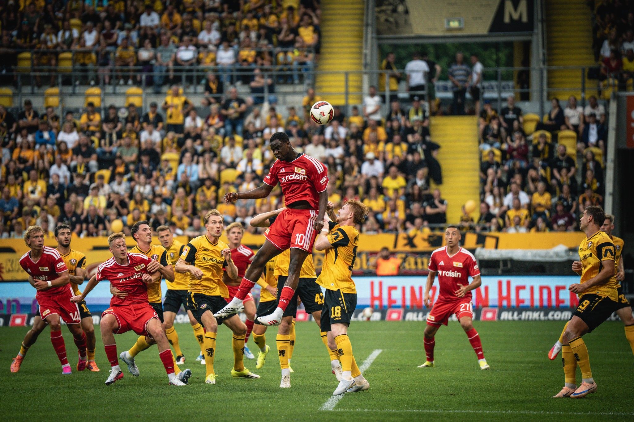 Spieler in roten Trikots springen zum Kopfball gegen Spieler in gelben Trikots vor einer leidenschaftlichen Menge im Stadion.