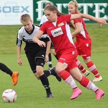 Ein Mädchen in rotem Fußballtrikot dribbelt den Ball, während ein Junge in schwarz-weißem Trikot sie verfolgt.