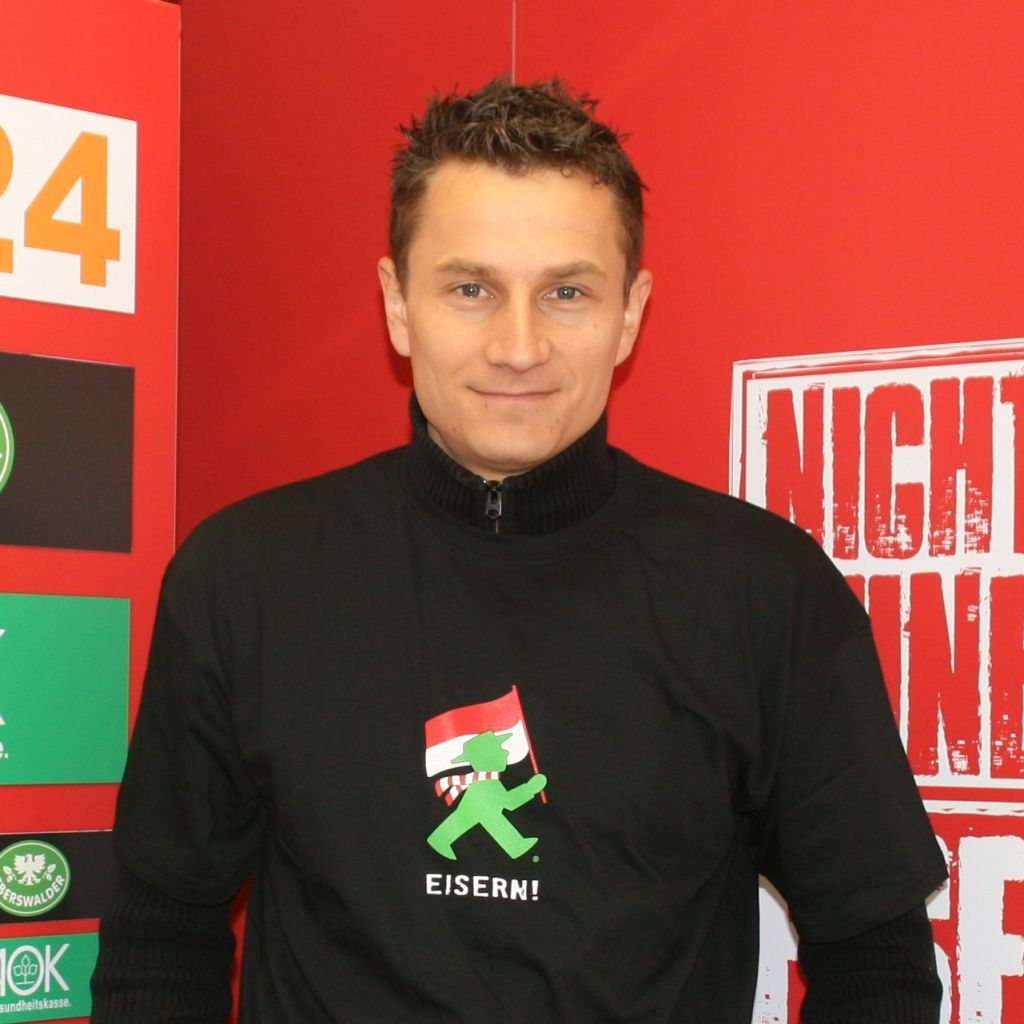 Mann mit kurzem, dunklem Haar trägt ein schwarzes T-Shirt mit dem Aufdruck „EISERN!“ und hält die nationale Flagge.