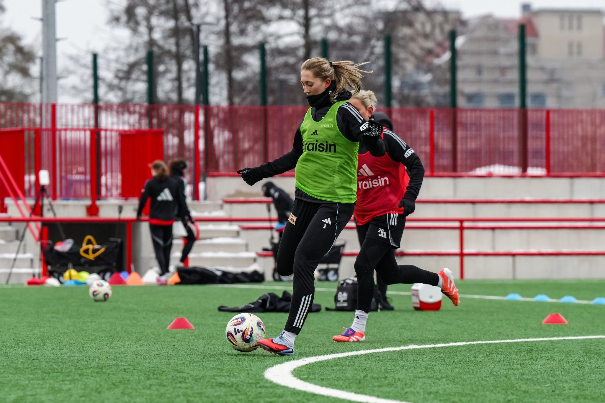 Zwei Fußballspielerinnen trainieren auf einem Kunstrasenplatz, eine trägt ein grünes Trainingsshirt, die andere rot.