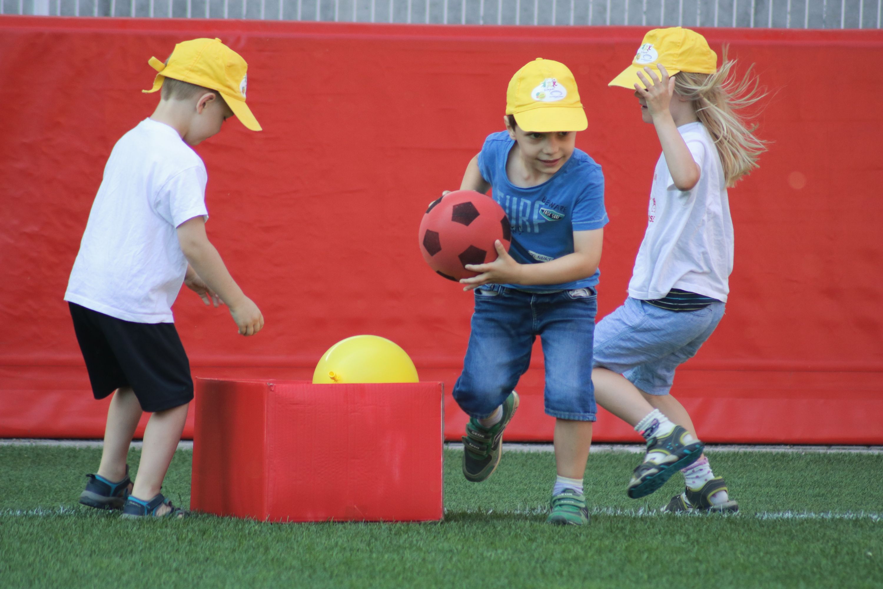 Drei Kinder in gelben Hüten spielen auf einem Rasenplatz mit einem roten Ball und einem gelben Ball, während eine rote Kiste im Hintergrund steht.