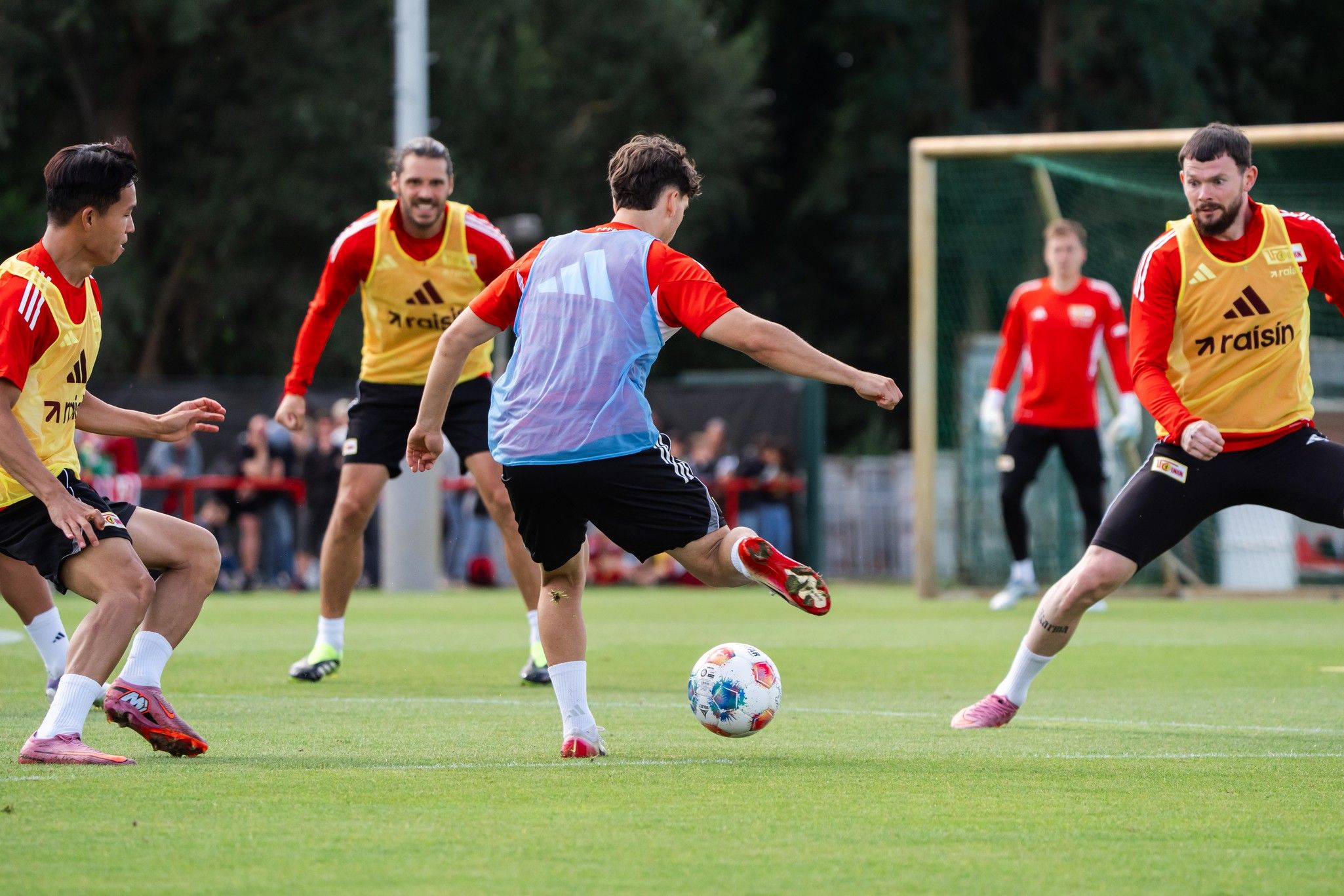 Fußballtraining mit mehreren Spielern auf dem Platz, ein Spieler mit Ball, andere beobachten und interagieren.