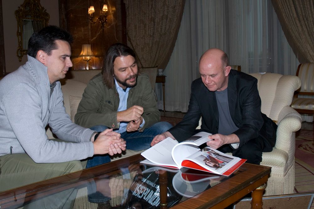 Drei Männer sitzen in einem eleganten Raum und betrachten ein Fotobuch auf einem Glastisch.