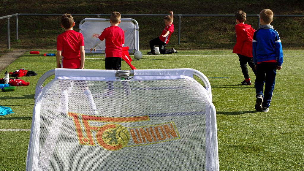Four children in red jerseys train on a grass field, with a soccer goal featuring the FC Union logo in the foreground.