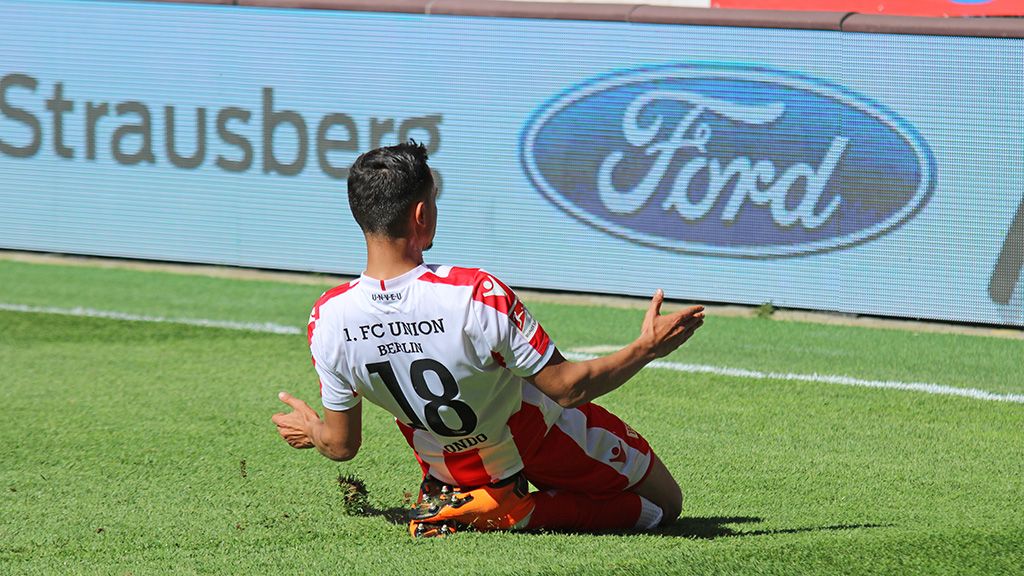 A soccer player from 1. FC Union Berlin slips on the grass after a play. Advertising banners in the background.