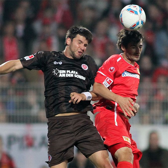 Zwei Fußballspieler in Aktion, einer mit braunem Trikot und der andere mit rotem Trikot, beim Kopfballduell.
