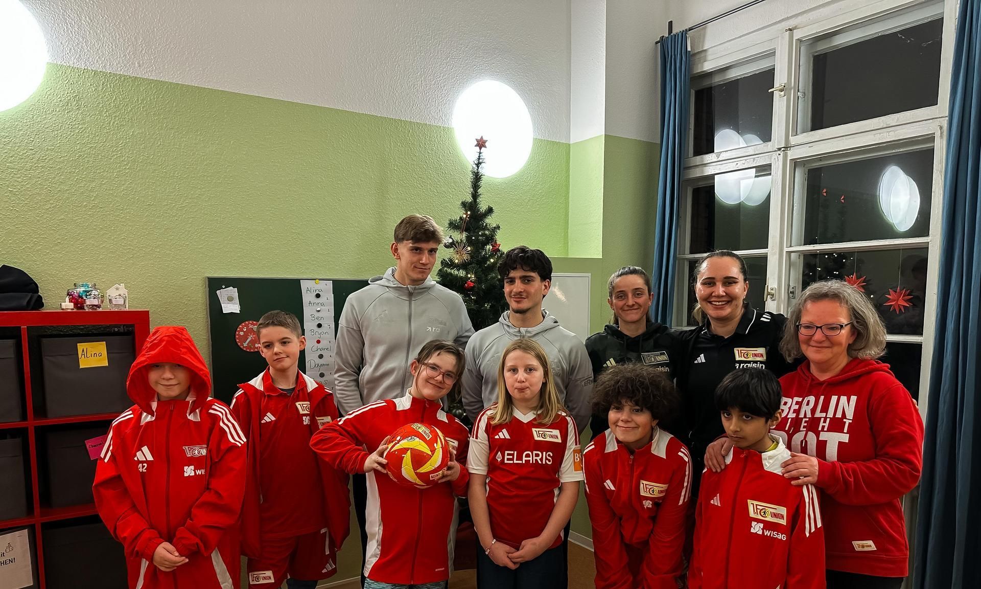 Eine Gruppe von Kindern und Jugendlichen in roten Sportanzügen steht vor einem Weihnachtsbaum in einem Raum.