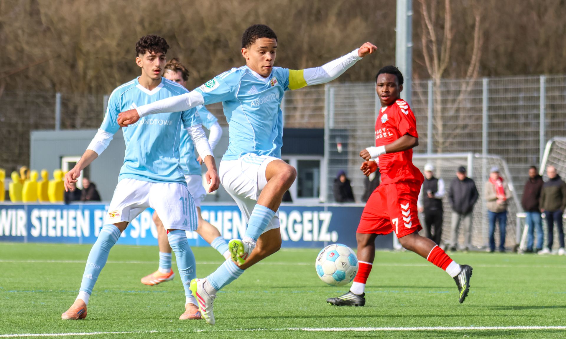 Ein Fußballspieler mit Kapitänsbinde schießt den Ball, während zwei Gegner in roten Trikots dem Spiel folgen.