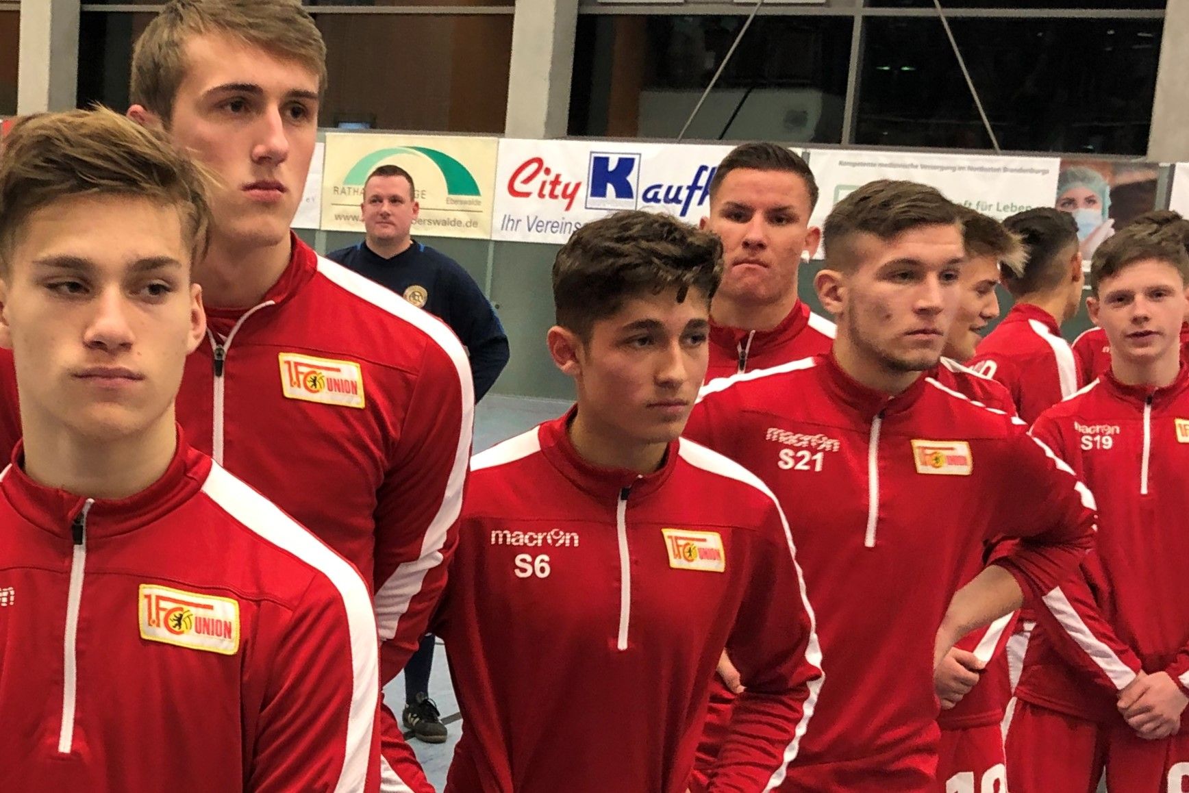 Gruppe junger Männer in roten Sportanzügen wartet auf den Beginn eines Wettkampfs in einer Sporthalle.