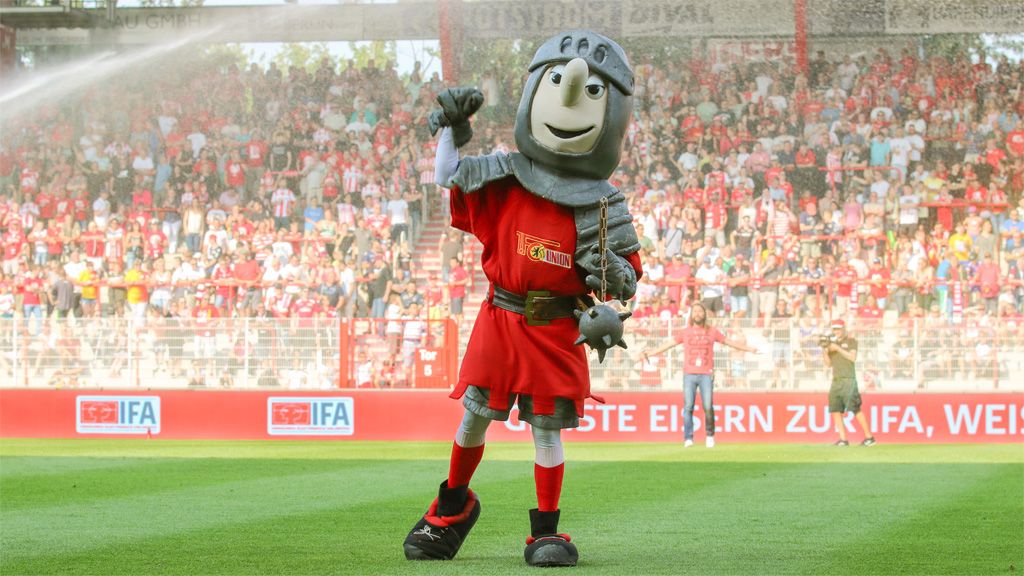 Ein Maskottchen in Ritterkostüm mit rot-grauer Kleidung, das auf einem Fußballfeld steht und jubelt, umgeben von Fans.