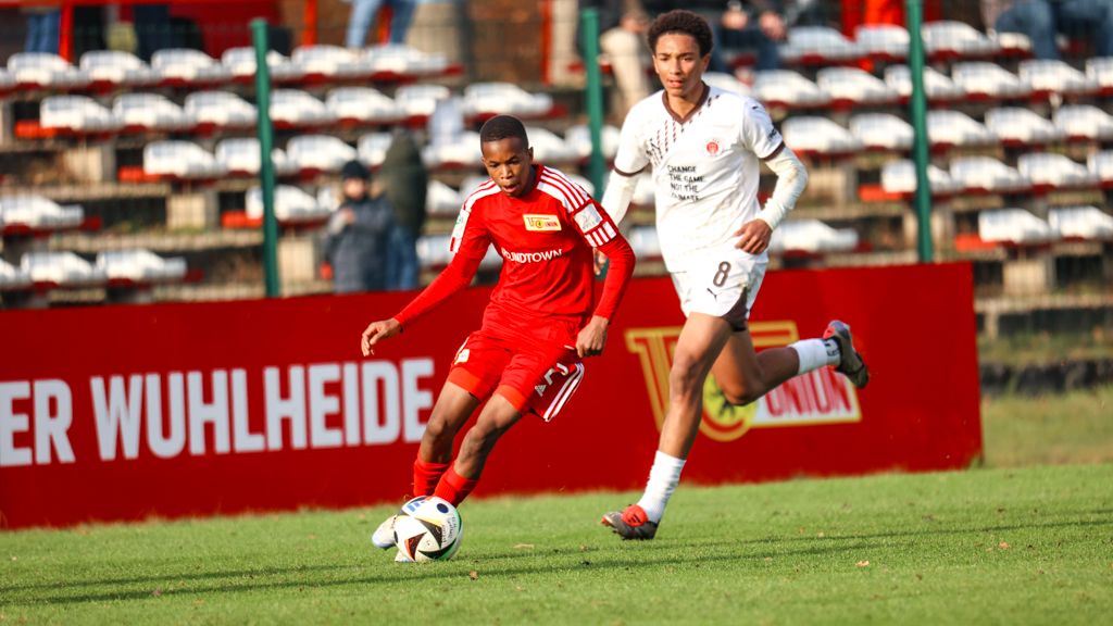 Ein junger Fußballspieler in rotem Trikot dribbelt den Ball, während ein gegnerischer Spieler in Weiß ihn verfolgt.