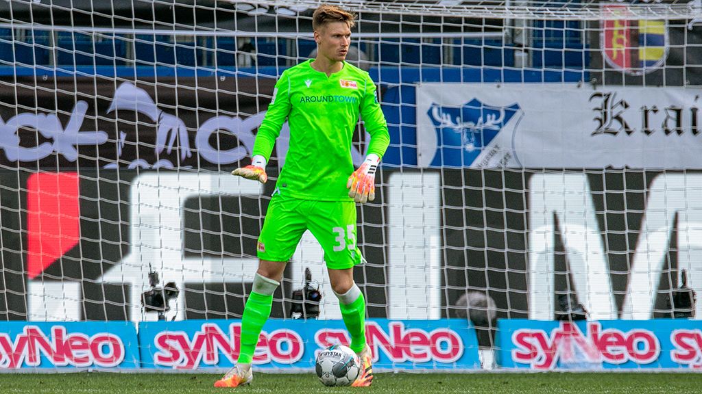 Torwart in neonfarbener Ausrüstung steht im Tor, bereit für den Spielzug, mit einem Fußballfeld im Hintergrund.
