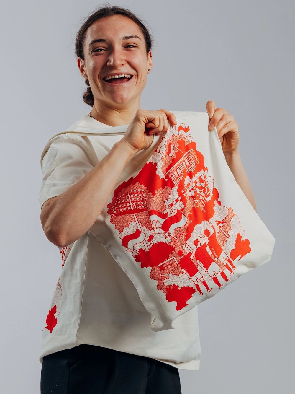 Frau mit einem großen, bedruckten Stoffbeutel in der Hand, lächelt fröhlich vor neutralem Hintergrund.