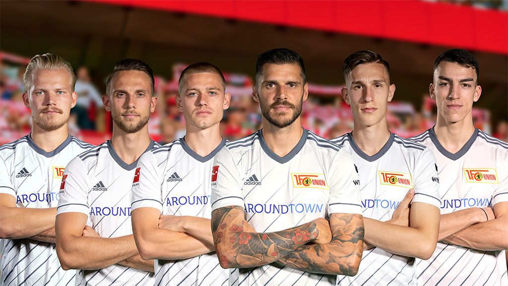 Sechs Fußballspieler in weißen Trikots stehen mit verschränkten Armen in Reihe, im Hintergrund eine unscharfe Stadionkulisse.